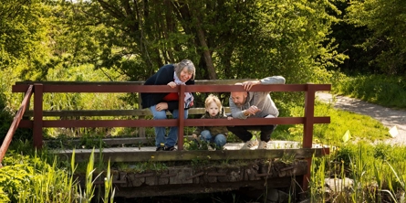 Billede af tre generationer på en lille bro over en lille å i naturen.