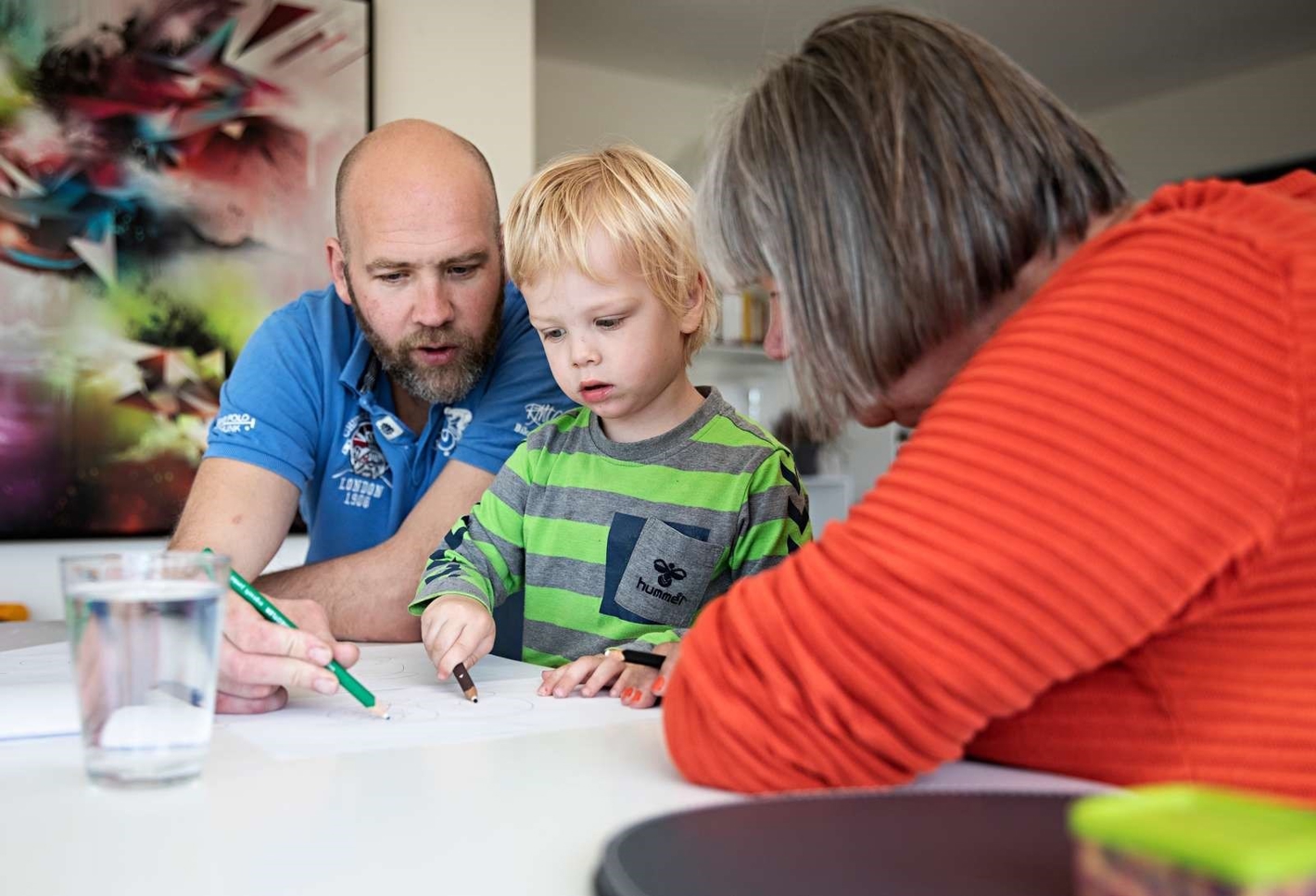 Billede af en ældre kvinde, en far og hans dreng, der sidder ved et bord og tegner.