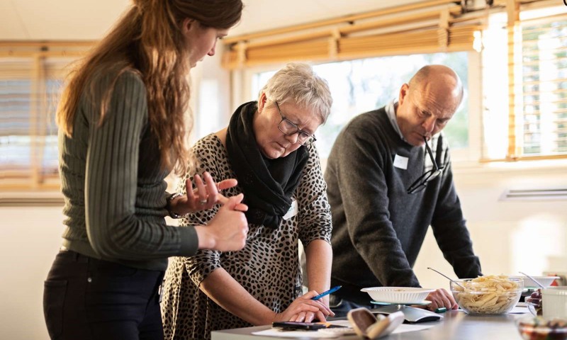 Billede af tre personer, der står ved et bord og lærer at tælle kulhydrater.